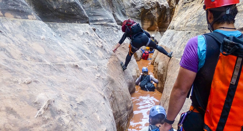 A group of people navigate through a narrow canyon filled with water. Some are wading in the waist-deep water, while another attempts to walk above the water, bracing their feet on either side of the canyon walls.
